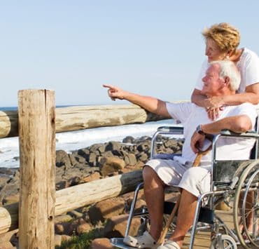 an elderly couple on a wooden boardwalk by the ocean. A woman with short blonde hair stands behind a man in a wheelchair