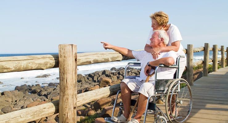 an elderly couple on a wooden boardwalk by the ocean. A woman with short blonde hair stands behind a man in a wheelchair