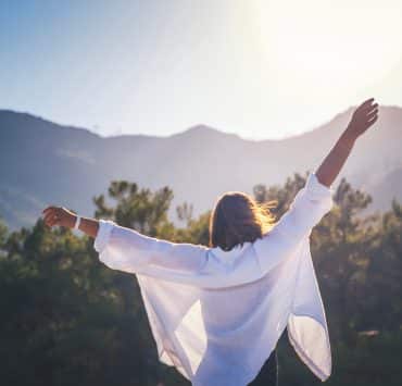 a woman from behind with her arms stretched wide against a scenic mountain backdrop