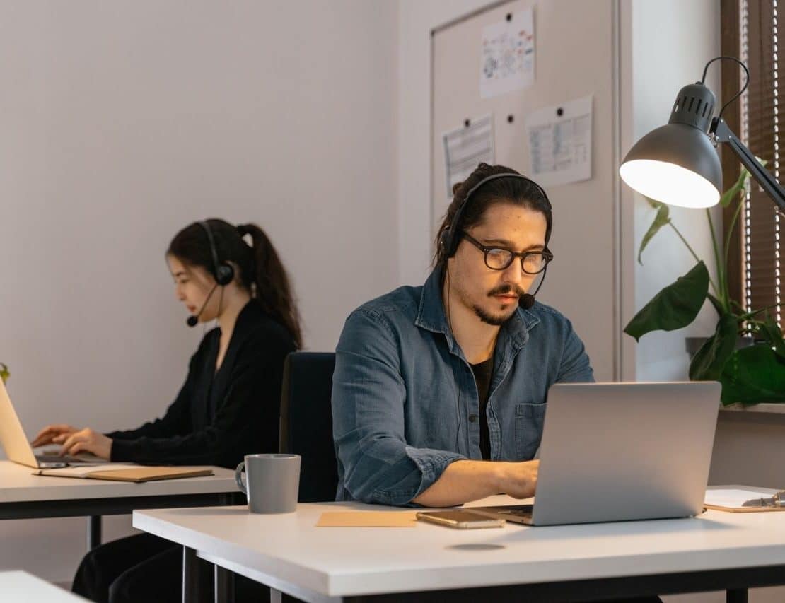 Two virtual assistants wearing headsets work on laptops in a modern office, providing remote administrative support.