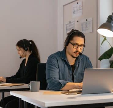 Two virtual assistants wearing headsets work on laptops in a modern office, providing remote administrative support.