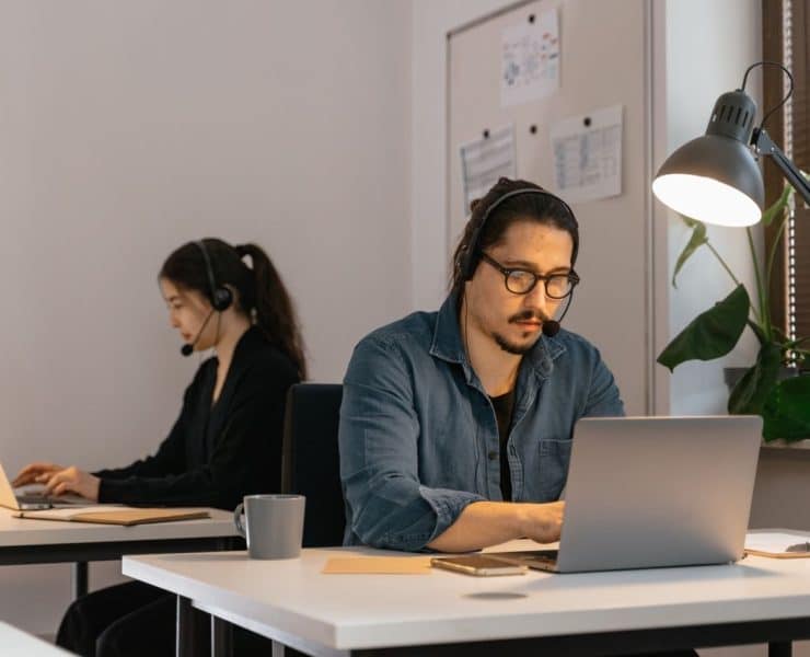 Two virtual assistants wearing headsets work on laptops in a modern office, providing remote administrative support.