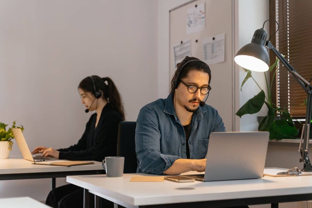Two virtual assistants wearing headsets work on laptops in a modern office, providing remote administrative support.