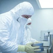two technicians wearing full white cleanroom hooded coveralls, face masks, and yellow protective gloves while handling metal trays in a sterile pharmaceutical or medical device manufacturing environment