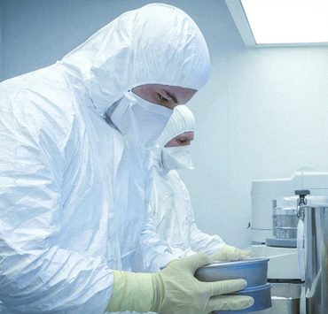 two technicians wearing full white cleanroom hooded coveralls, face masks, and yellow protective gloves while handling metal trays in a sterile pharmaceutical or medical device manufacturing environment