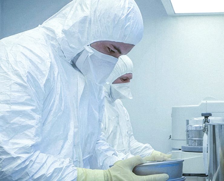 two technicians wearing full white cleanroom hooded coveralls, face masks, and yellow protective gloves while handling metal trays in a sterile pharmaceutical or medical device manufacturing environment