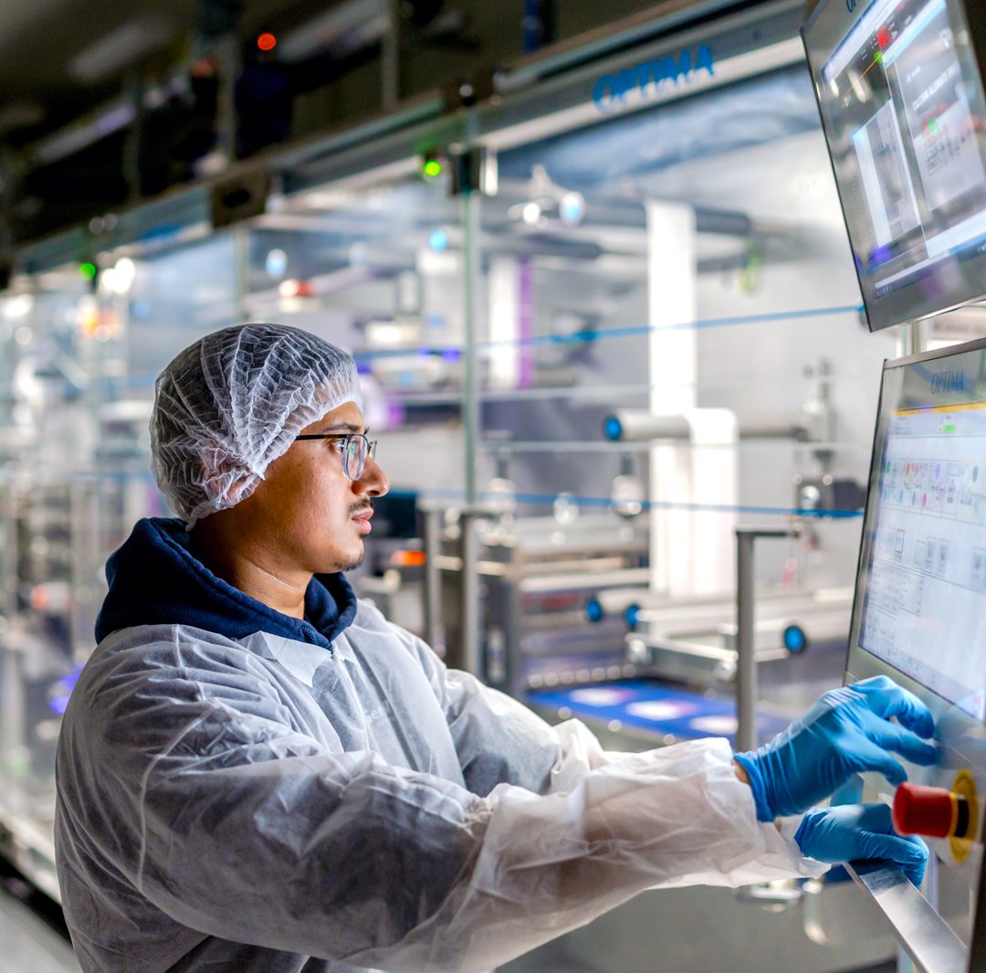 worker in cleanroom personal protective equipment including a hairnet, face mask, lab coat, and blue nitrile gloves—operating an automated industrial touchscreen control panel in a manufacturing or processing facility