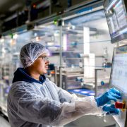 worker in cleanroom personal protective equipment including a hairnet, face mask, lab coat, and blue nitrile gloves—operating an automated industrial touchscreen control panel in a manufacturing or processing facility