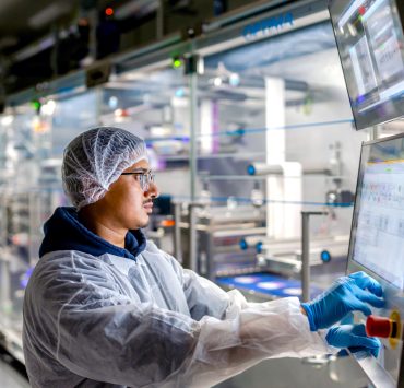 worker in cleanroom personal protective equipment including a hairnet, face mask, lab coat, and blue nitrile gloves—operating an automated industrial touchscreen control panel in a manufacturing or processing facility