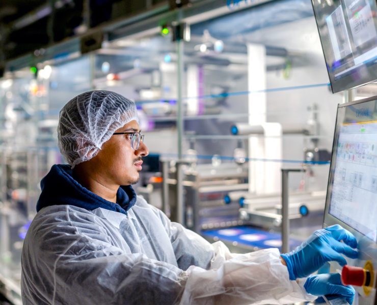 worker in cleanroom personal protective equipment including a hairnet, face mask, lab coat, and blue nitrile gloves—operating an automated industrial touchscreen control panel in a manufacturing or processing facility