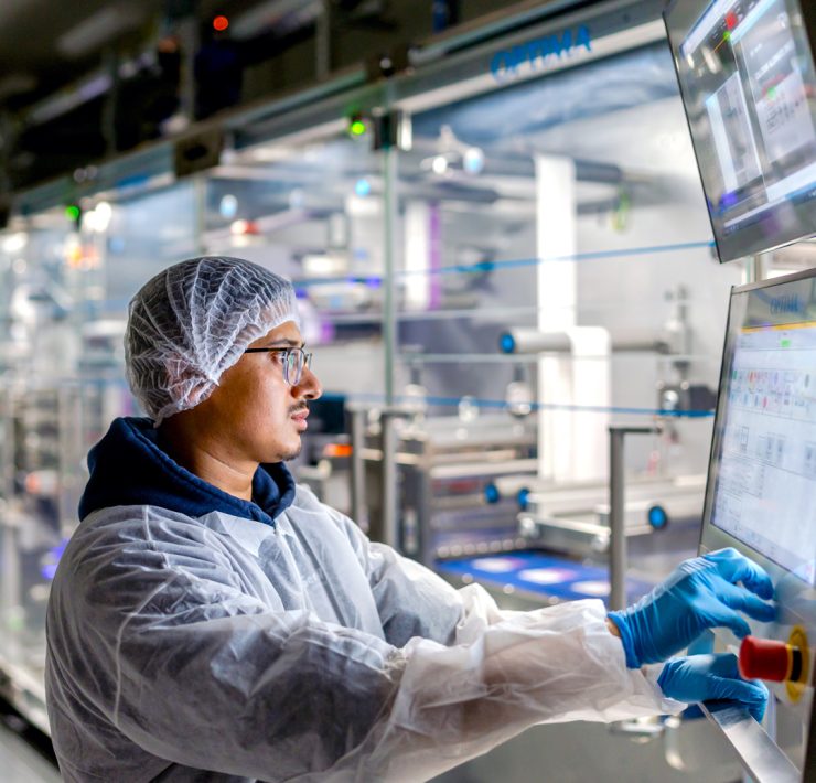 worker in cleanroom personal protective equipment including a hairnet, face mask, lab coat, and blue nitrile gloves—operating an automated industrial touchscreen control panel in a manufacturing or processing facility