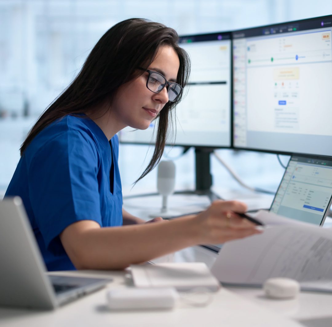 healthcare technician or administrative worker reviewing medical records and billing codes on a laptop and multiple monitors