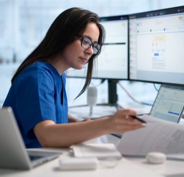healthcare technician or administrative worker reviewing medical records and billing codes on a laptop and multiple monitors
