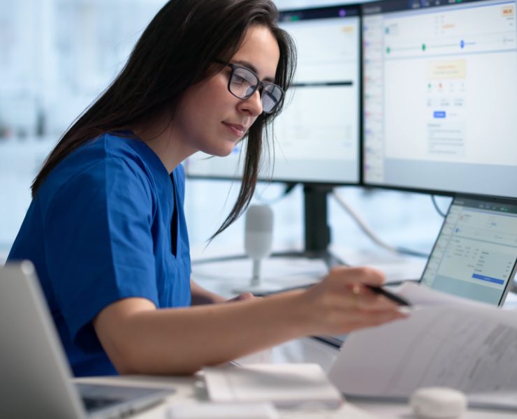 healthcare technician or administrative worker reviewing medical records and billing codes on a laptop and multiple monitors